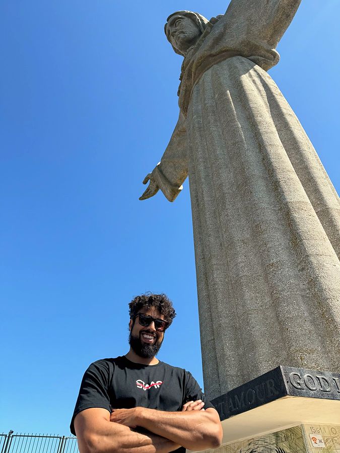A man in a crossed-out SIMP tee stands beneath the Cristo Rei statue