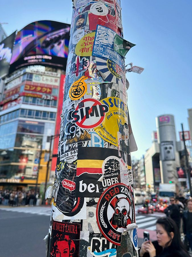 A sticker-covered pole at Shibuya crossing with the NO SIMP sticker