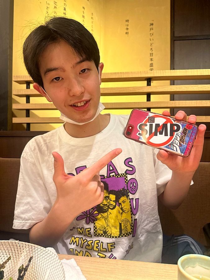 A young man in Tokyo points at the NO SIMP sticker on his phone case