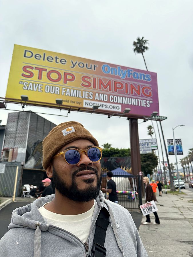 A marcher in a beanie and sunglasses stands beneath the Delete Your OnlyFans billboard